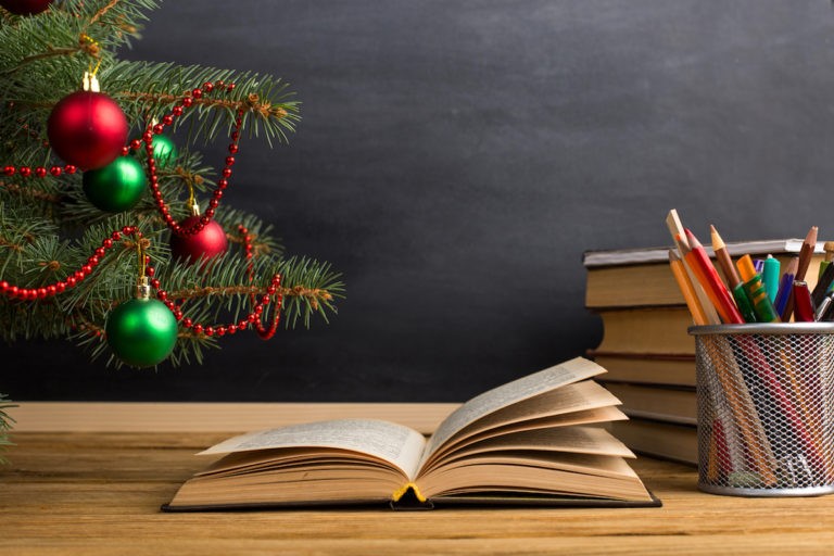Open book on a desk next to a Christmas tree, stack of books, and cup of pencils.