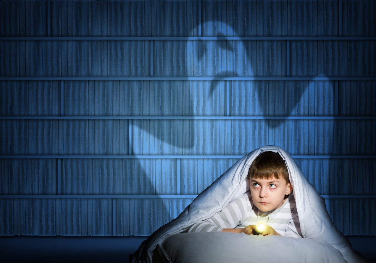 Boy under blankets with flashlight while ghost is on the wall behind him