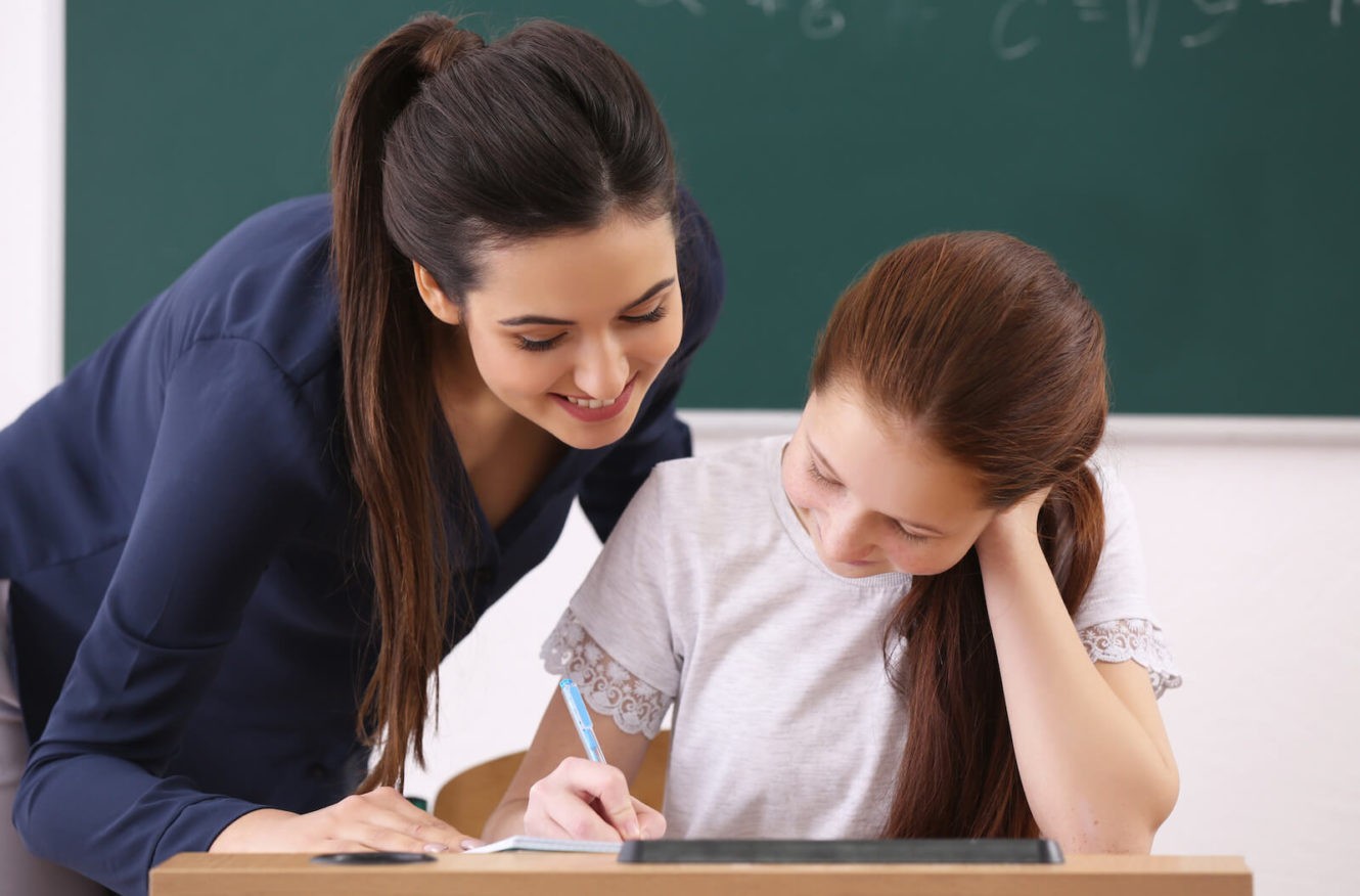 Female teacher helping young girl with school work at her desk