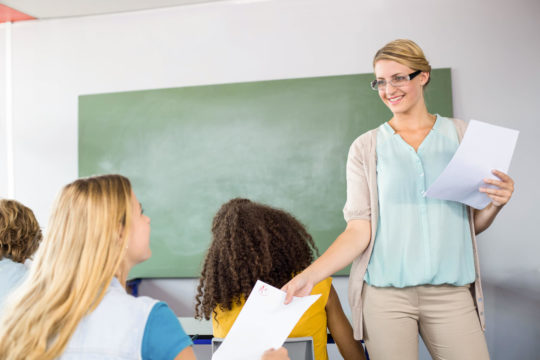 Female teacher handing a paper back to a seated student in a classroom.
