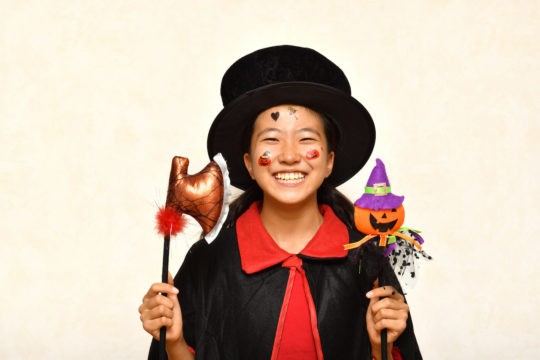A girl dressed in a costume with a top hat smiles at the camera, holding a Halloween decoration.