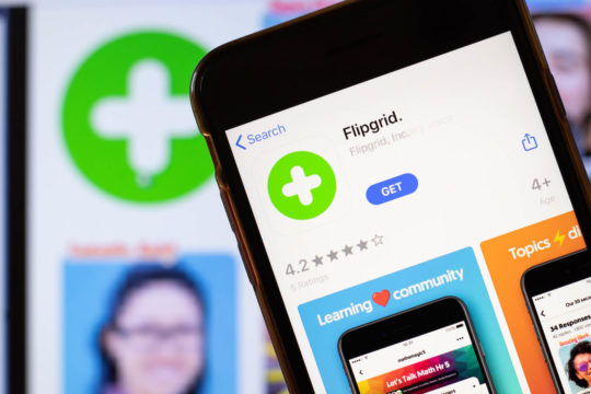 A close up of the Flipgrid app logo on a phone screen, with the Flipgrid website on a computer in the background.