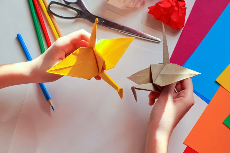 Children鈥檚 hands make an origami crane, above a white background with various school supplies.