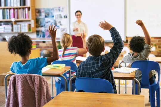 A classroom of students raise their hands as their teacher asks them a question.