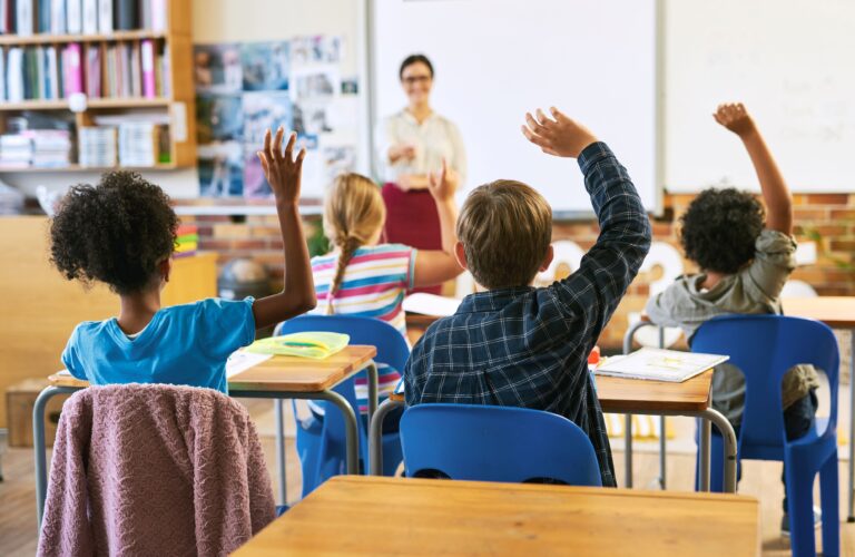 A classroom of students raise their hands as their teacher asks them a question.