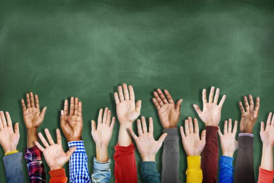 A close-up of student hands being raised in front of a chalkboard, representing student participation.